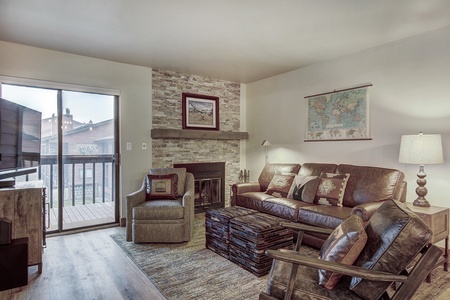 Cozy living room featuring a leather sofa, armchair, wooden rocking chair, fireplace, wall maps, sliding glass door to balcony, and wooden furniture. Light from the lamp and balcony brightens the space.