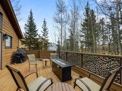 A wooden deck with patio furniture, a table, and a barbecue grill surrounded by trees and a forested area in the background.