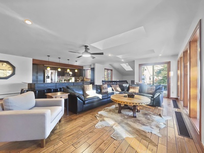 Open-concept living room featuring a black leather sectional, light grey armchair, round wooden coffee table, cowhide rug, and a kitchen in the background with pendant lights and bar stools.