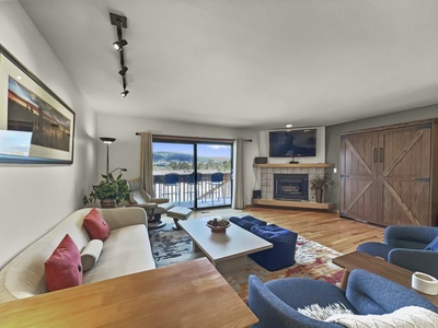 A cozy living room features a beige couch with red pillows, a flat-screen TV, a fireplace, a sliding glass door opening to a balcony, and a view of a mountainous landscape in the background.