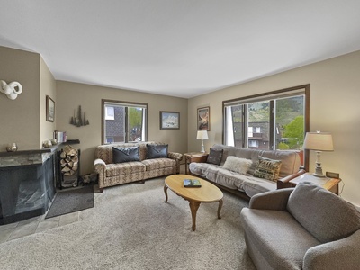 A cozy living room with two sofas, an armchair, a coffee table, and a fireplace. Large windows provide natural light and offer views of greenery outside. The room features neutral tones and carpet flooring.