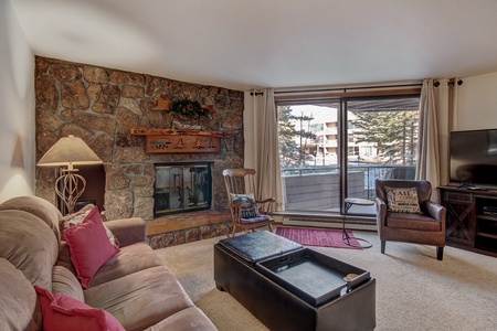 A cozy living room with a stone fireplace, a plush sofa with red pillows, a rocking chair, and a coffee table. A large window shows a snowy outdoor scene. A TV stands on the right side.