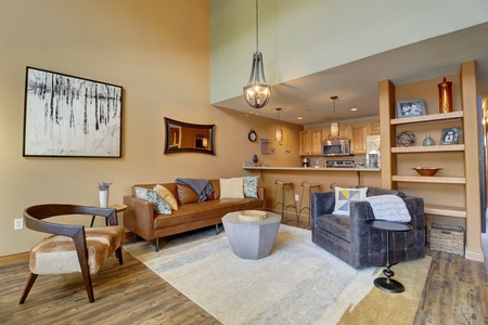 A modern living room with a brown sofa, armchair, and a round coffee table. The room features a two-story ceiling, a large chandelier, wall art, and an open view into a kitchenette area.