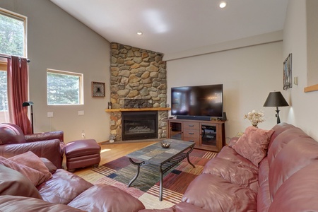 A living room with a red leather sectional sofa, stone fireplace, large flat-screen TV, and a coffee table on a multicolored rug, with natural light coming in from window.