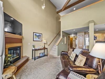 A cozy living room with a sectional sofa, a fireplace, a flatscreen TV above the mantle, and a staircase leading to the upper floor. The room features warm lighting and a view into the kitchen area.