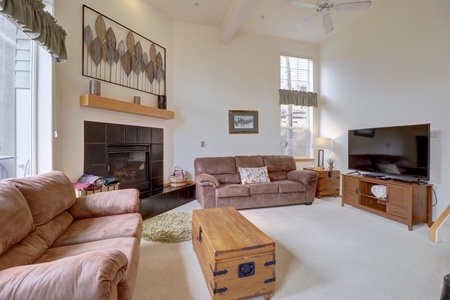 A living room with two brown leather sofas, a wooden chest as a coffee table, a fireplace, and a large TV on a stand. The room has high ceilings, a ceiling fan, and wall art above the fireplace.