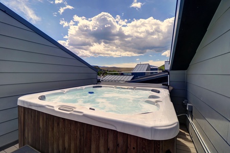 Outdoor hot tub on a rooftop deck between two sloped roofs, with a clear blue sky and scattered clouds in the background.