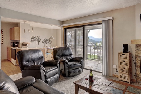 A living area with leather recliners, a coffee table with wine glasses and a bottle, a sliding glass door opening to a balcony, and a kitchen in the background.