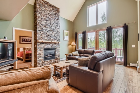 Living room with high ceilings, stone fireplace, brown leather sofas, and large windows next to a sliding door leading to an outdoor balcony. Bedroom visible through doorway on the left.