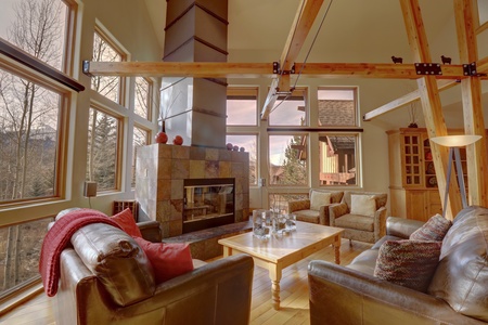 A cozy living room with large windows, two leather armchairs, a fabric sofa, a stone fireplace, and a wooden coffee table. Exposed wooden beams and trees are visible through the windows.