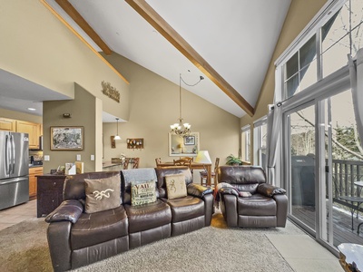 A spacious, well-lit living room with high ceilings, large windows, a leather sofa and recliner, a dining area in the background, and a view of trees outside.