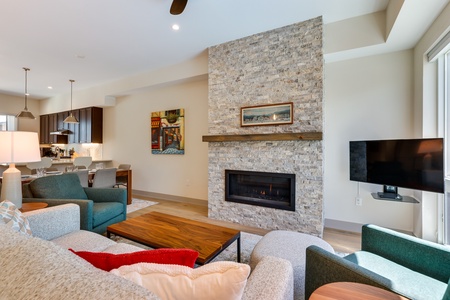 A modern living room with a stone fireplace, wall-mounted TV, and colorful artwork. Light grey sofas and teal chairs are arranged around a wooden coffee table. Dining area and kitchen visible in background.