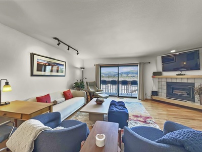 A bright living room with white walls, featuring a cozy sofa, armchairs, coffee table, wall art, track lighting, a fireplace, TV, and large sliding doors opening to a balcony with a mountain view.