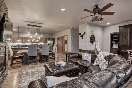 A spacious open-plan living room and kitchen with wooden floors, leather sectional sofa, ceiling fan, modern light fixtures, and dark wood cabinetry.