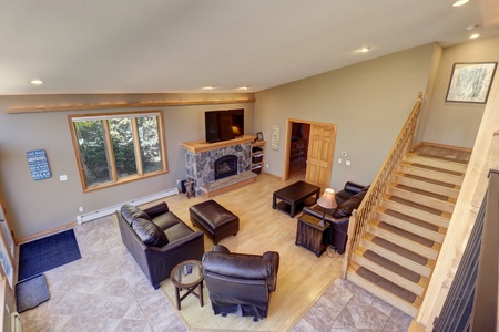 Spacious living room with a stone fireplace, leather seating, wooden coffee table, and large window. A staircase leads to an upper level. Walls are painted beige and the room has a mix of tile and wood flooring.