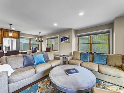 A modern living room with beige sofas and blue pillows, a large round coffee table, and a view of the dining area and kitchen in the background. Multiple windows let in natural light.