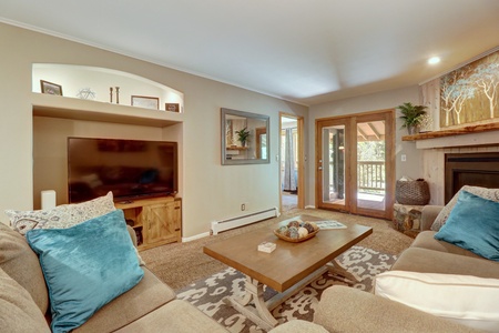 A cozy living room with a wall-mounted TV, neutral-toned sofas with blue pillows, a wooden coffee table, and glass doors leading to a balcony. Shelves and a mirror decorate the wall above the TV.