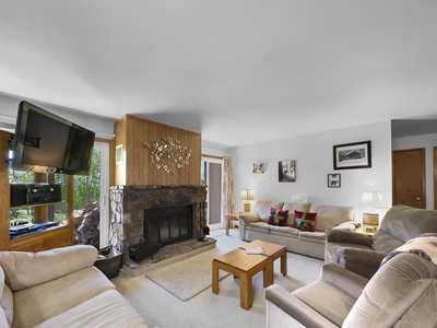 A cozy living room with a stone fireplace, a TV mounted on the wall, multiple seats, wooden furniture, and decorated with framed photos.