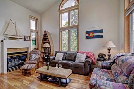 A living room with tall windows, a brown leather sofa, a patterned couch, a fireplace, a bookshelf, and a boat model. A coffee table with wine glasses and a bottle sits in the center.