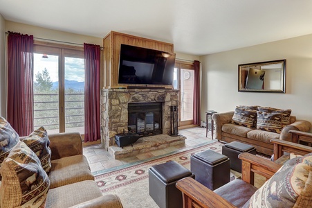 A cozy living room with stone fireplace, mounted TV, plush seating, dark wood furniture, and large windows with red curtains overlooking a scenic view. A large mirror hangs on the right wall.