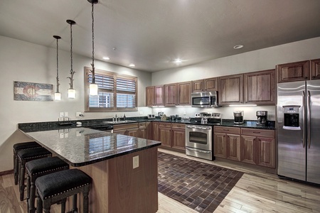 A modern kitchen with an island and three bar stools. It features wooden cabinets, stainless steel appliances, and pendant lighting. A rug lies between the island and cabinets.