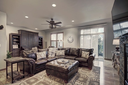 A cozy living room featuring a large leather sectional sofa, tufted ottoman, and a fireplace. Wooden furniture and decor items complement the space, which includes a large window with a scenic view.