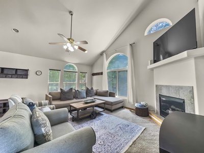 A spacious, well-lit living room with large windows, neutral-colored furniture, a ceiling fan, a fireplace, and a flat-screen TV mounted above the mantel.