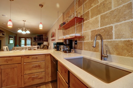 A modern kitchen features a large sink, wooden cabinets, open shelving with glassware, and a stone backsplash. Hanging pendant lights illuminate the countertop area. Dining room visible in the background.