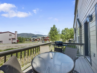 A glass-topped table with chairs on a wooden deck overlooks a rural street, houses, trees, and distant hills under a blue sky with scattered clouds. A grill stands near the railing.