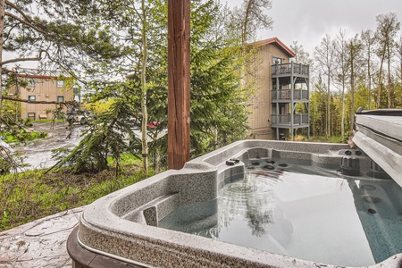 Outdoor hot tub with water filled to the top, surrounded by a wooded area and adjacent to an apartment building. Overcast sky and wet pavement in the background.