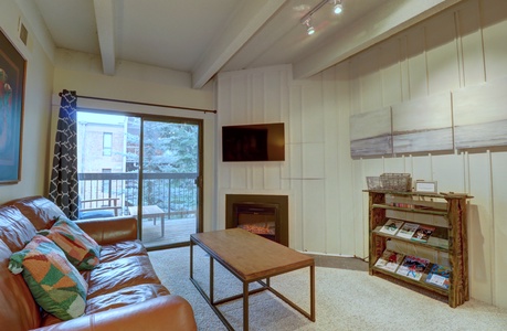 Living room with a leather sofa, wooden coffee table, and a wall-mounted TV above an electric fireplace. A bookcase with magazines is on the right. Sliding glass doors lead to a balcony.