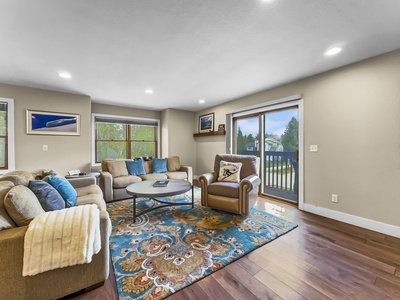 A cozy living room with a large area rug, sofas, an armchair, a coffee table, large windows, and a sliding glass door leading to a deck with a view of trees.