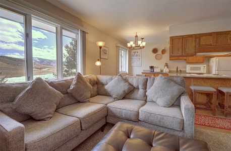 A cozy living room with a large sectional sofa, a leather ottoman, and large windows overlooking a mountainous landscape. Adjacent is a kitchen with wooden cabinets and a breakfast bar with stools.