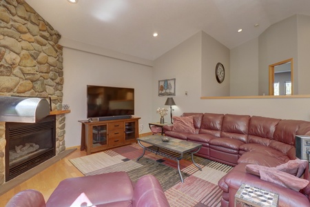 A living room with a large red leather sectional sofa, a stone fireplace, a flat-screen TV on a wooden stand, and a glass coffee table on a patterned rug. Light-colored walls and recessed lighting.