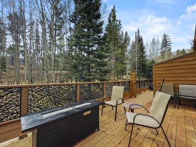 Wooden deck with two chairs and a rectangular fire pit table, overlooking a forested area. Decorative railing panels add design to the deck. Cloudy sky in the background.