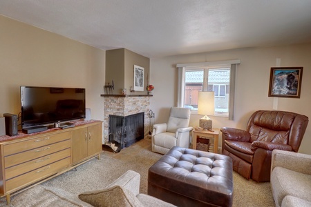 A cozy living room with a TV on a wooden stand, a fireplace, an armchair, a recliner, a window with blinds, and a framed picture on the wall. A brown ottoman sits in the center.