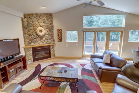 Living room with a stone fireplace, leather sofas, a wall-mounted TV, and a coffee table on a colorful rug. Large windows and glass doors lead to an outdoor deck with trees visible outside.