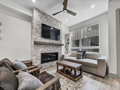 Modern living room with stone fireplace, mounted TV, ceiling fan, and large window. Furnishings include a gray couch, two wooden armchairs with gray cushions, and a wooden coffee table.