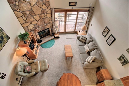 Bird's-eye view of a cozy living room featuring a stone fireplace, gray sofa, armchair, wooden coffee table, and a sliding glass door leading to a balcony with seating. Walls are adorned with decor.