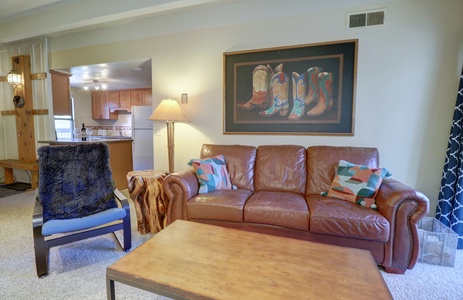 A living room with a brown leather sofa, colorful pillows, wooden coffee table, cushioned chair, lamp, and a framed painting of cowboy boots on the wall. Kitchen area visible in the background.