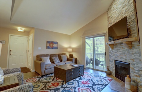 A cozy living room features a stone fireplace, flat-screen TV, patterned rug, beige sofa, armchair, coffee table, and sliding glass doors leading to a balcony with a scenic view.