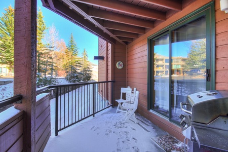 A balcony with snowy floor, white Adirondack chairs, a small table, and a barbecue grill. The wooden railing overlooks a scenic view of snow-covered trees and nearby buildings.