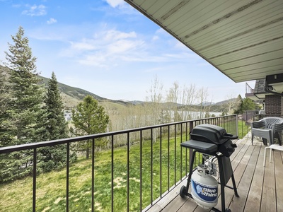 A balcony with a gas grill overlooks a scenic view of trees, a grassy area, and distant rolling hills under a partly cloudy blue sky.