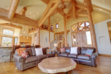 A spacious living room with high wooden beams and large windows, featuring a brown leather sectional, a rustic round coffee table, and various cushions. The room opens to a kitchen and a staircase.