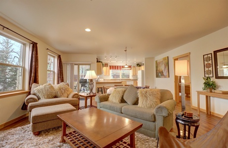 A cozy living room featuring a large window with brown curtains, a gray sofa with pillows, an armchair, a wooden coffee table, and a view of the dining area and kitchen in the background.