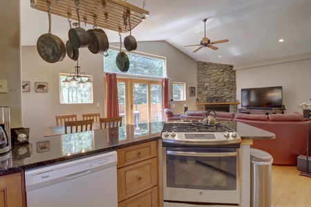 Open-concept kitchen and living room with a granite countertop, hanging pots, wooden cabinets, stainless steel stove, dishwasher, ceiling fan, stone fireplace, television, and sliding glass doors.