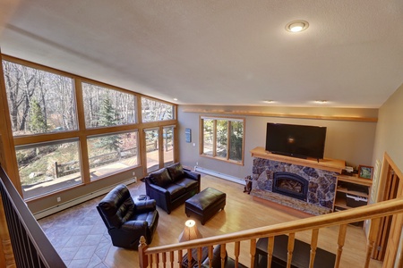 A living room with large windows, a hardwood floor, leather furniture, a stone fireplace, and a flat-screen TV. The outside view shows trees and a spacious wooden deck. Stairs lead down into the room.