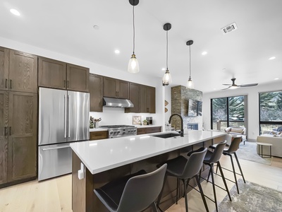 Modern kitchen with stainless steel appliances, dark wood cabinets, and a large island with black stools. Pendant lights hang above the island, and the kitchen opens to a living area with large windows.