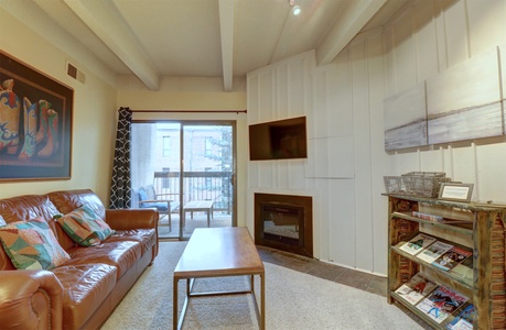 A living room features a brown leather sofa, a wooden coffee table, a fireplace, a wall-mounted TV, a bookshelf with magazines, and a large painting. A window with a sliding door opens to a balcony.