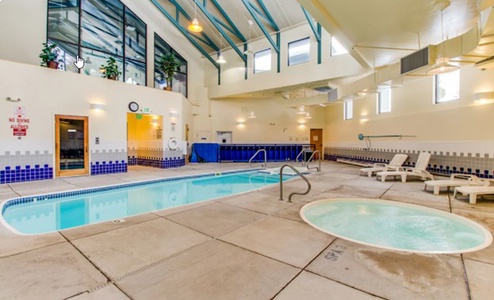 Indoor swimming pool area with a small hot tub, lounge chairs, windows for natural light, and a high ceiling. Lifesaving equipment is mounted on the walls.